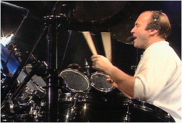 Phil Collins singing into a headset microphone while playing drums in a live performance.